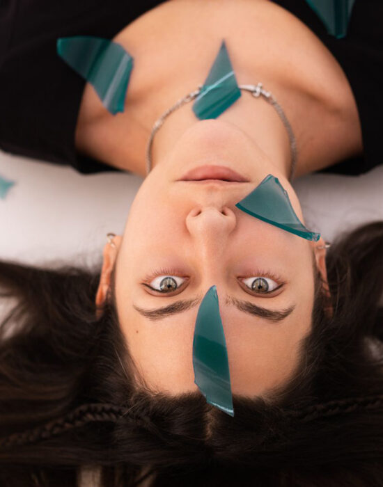 Fotografia a mezzo busto di una donna giovane, distesa su una superficie, i suoi capelli sono castani e i suoi occhi sono chiari. Guarda fissa sull'obiettivo e dei coriandoli blu semi-trasparenti le cadono sul volto.
